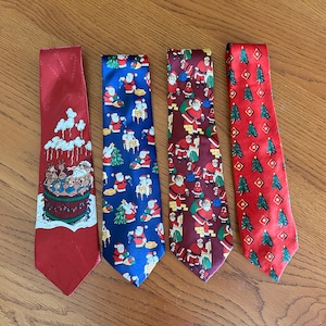 Puede incluir: Cuatro corbatas con temática navideña. Las corbatas presentan diseños festivos, incluyendo a Papá Noel, árboles de Navidad y renos. Las corbatas son rojas, azules y burdeos, y están dispuestas sobre una superficie de madera.