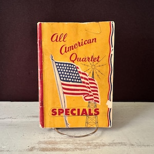 Könnte beinhalten: Ein Vintage-Buch mit dem Titel "All American Quartet Specials" mit einer Illustration der amerikanischen Flagge. Das Cover ist gelb mit roten und blauen Akzenten. Das Buch wird auf einem Metallständer präsentiert.