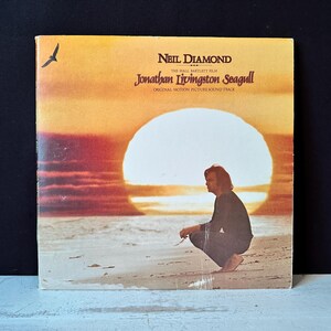 Peut inclure: Pochette d'album vinyle vintage pour la bande originale de "Jonathan Livingston Seagull" de Neil Diamond. La couverture présente une scène de coucher de soleil avec une personne sur la plage et le titre de l'album. Une mouette vole dans le coin supérieur gauche.