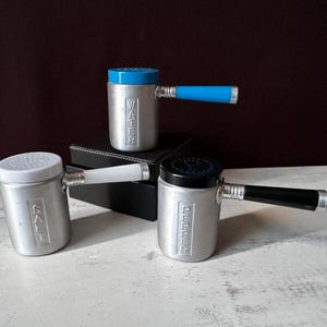 Könnte beinhalten: Drei silberne Streuer mit schwarzen, weißen und blauen Deckeln. Die Streuer haben Griffe und sind mit "SALT", "WATER" und "PEPPER" beschriftet.