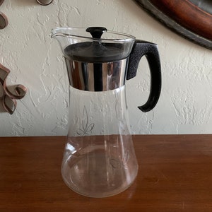 Può includere: Una caraffa di vetro trasparente con un coperchio e un manico neri. La caraffa ha una fascia argentata attorno alla parte superiore e un motivo decorativo sul lato.