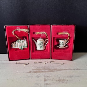 May include: Three porcelain Christmas ornaments featuring a mailbox, a teapot, and a teacup with saucer, each decorated with green holly leaves and red berries. The ornaments are housed in individual red velvet-lined boxes with gold hanging cords.