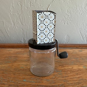 May include: A vintage hand-crank food grinder with a white and black geometric pattern. The grinder has a clear glass jar and a black plastic lid.