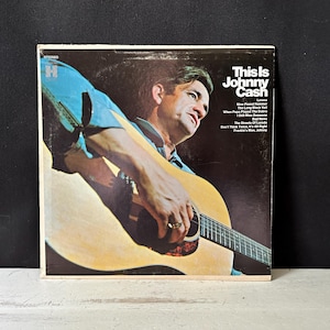 May include: Vintage vinyl record album cover featuring Johnny Cash playing a guitar. The album is titled "This Is Johnny Cash" and includes a list of songs. The cover art shows Cash in a blue shirt.