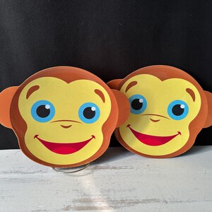 Puede incluir: Dos decoraciones con forma de mono con orejas marrones, caras amarillas, ojos azules y sonrisas rojas. Las caras de mono están sobre una superficie blanca con un fondo negro. Las decoraciones son probablemente para la habitación de un niño o una fiesta.