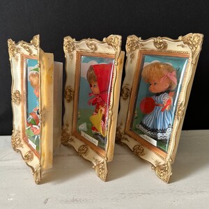 May include: Three miniature doll picture frames with gold trim. Each frame features a different doll with a different outfit and a different name on the bottom. The dolls are dressed in a red and white dress, a blue and white dress, and a green and white dress.