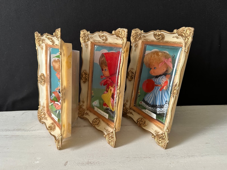 May include: Three miniature doll picture frames with gold trim. Each frame features a different doll with a different outfit and a different name on the bottom. The dolls are dressed in a red and white dress, a blue and white dress, and a green and white dress.