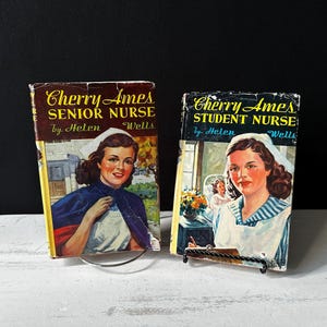 May include: Two vintage paperback books with covers featuring illustrations of nurses. The book on the left is titled "Cherry Ames Senior Nurse" by Helen Wells. The book on the right is titled "Cherry Ames Student Nurse" by Helen Wells.