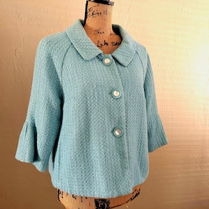 Könnte beinhalten: Hellblaue Jacke mit drei weißen Knöpfen und einem runden Kragen. Die Jacke hat dreiviertellange Ärmel mit ausgestelltem Design. Der Stoff hat ein strukturiertes Aussehen. Die Jacke wird auf einer Schneiderpuppe präsentiert.