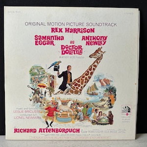 Puede incluir: Una portada de álbum de vinilo vintage para la película Doctor Dolittle. La portada presenta a Rex Harrison, Samantha Eggar y Anthony Newley. La portada del álbum es un collage vibrante de imágenes de la película, que incluye una jirafa, un loro y una variedad de otros animales.