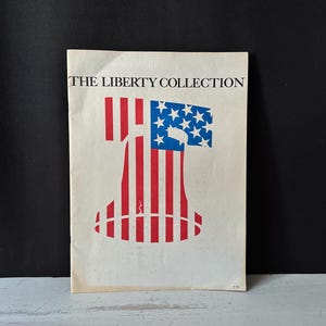 May include: A vintage print titled "THE LIBERTY COLLECTION" featuring a stylized illustration of the Statue of Liberty. The design incorporates the American flag with red and white stripes and a blue field of stars. The print is on a cream-colored paper.