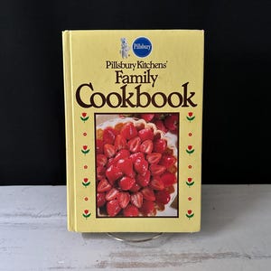 May include: A vintage Pillsbury Kitchens Family Cookbook with a yellow cover. The cover features the Pillsbury logo and a picture of a strawberry pie. Red and green floral accents decorate the edges of the book.