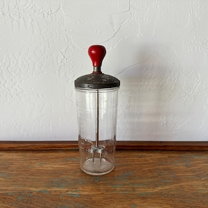 Könnte beinhalten: Ein Vintage-Cocktailmixer aus Glas mit einem Metalldeckel und einem roten Holzknauf. Das transparente Glasgefäß hat Messmarkierungen und einen Schneebesen aus Metall im Inneren. Der Mixer steht auf einer Holzoberfläche.