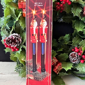 Puede incluir: Una caja de cartón rectangular con la imagen de dos velas cónicas con forma de cascanueces. La caja es roja con bordes dorados y muestra el texto SET OF 2 10" Tapers. Las velas tienen detalles en colores rojo, azul y negro.