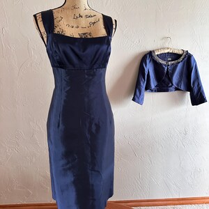 May include: A navy blue formal dress with a square neckline and a matching bolero jacket. The dress is displayed on a mannequin, and the bolero is hanging on a hanger. The dress is knee-length.