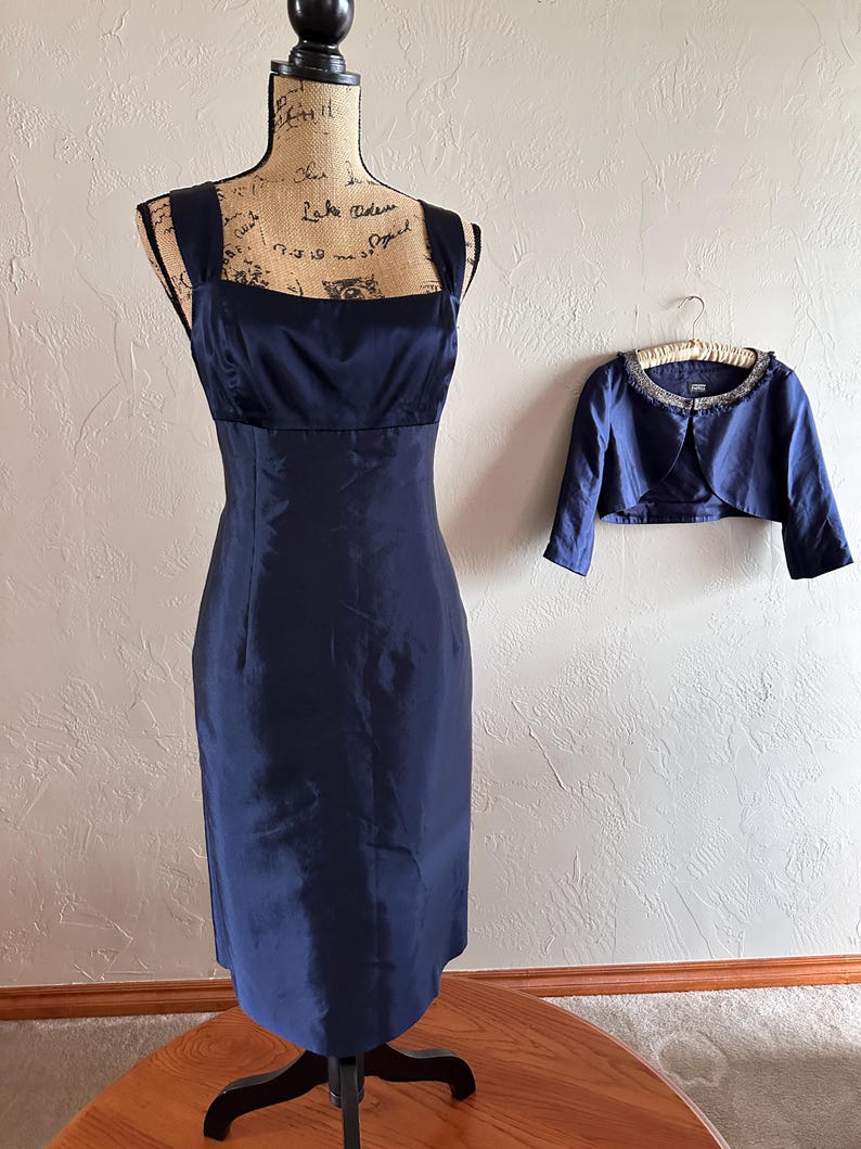 May include: A navy blue formal dress with a square neckline and a matching bolero jacket. The dress is displayed on a mannequin, and the bolero is hanging on a hanger. The dress is knee-length.