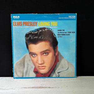 Peut inclure: Pochette d'album vintage d'Elvis Presley, "Loving You". La couverture présente un portrait d'Elvis en veste grise et chemise rouge sur fond bleu. Le titre de l'album et les titres des chansons sont visibles.