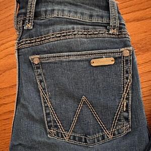 Pode incluir: Close-up do bolso traseiro de jeans azuis. O bolso apresenta um design de costura branco distinto e uma pequena etiqueta retangular com a marca "Wrangler". Os jeans t&ecirc;m um visual cl&aacute;ssico e desgastado.