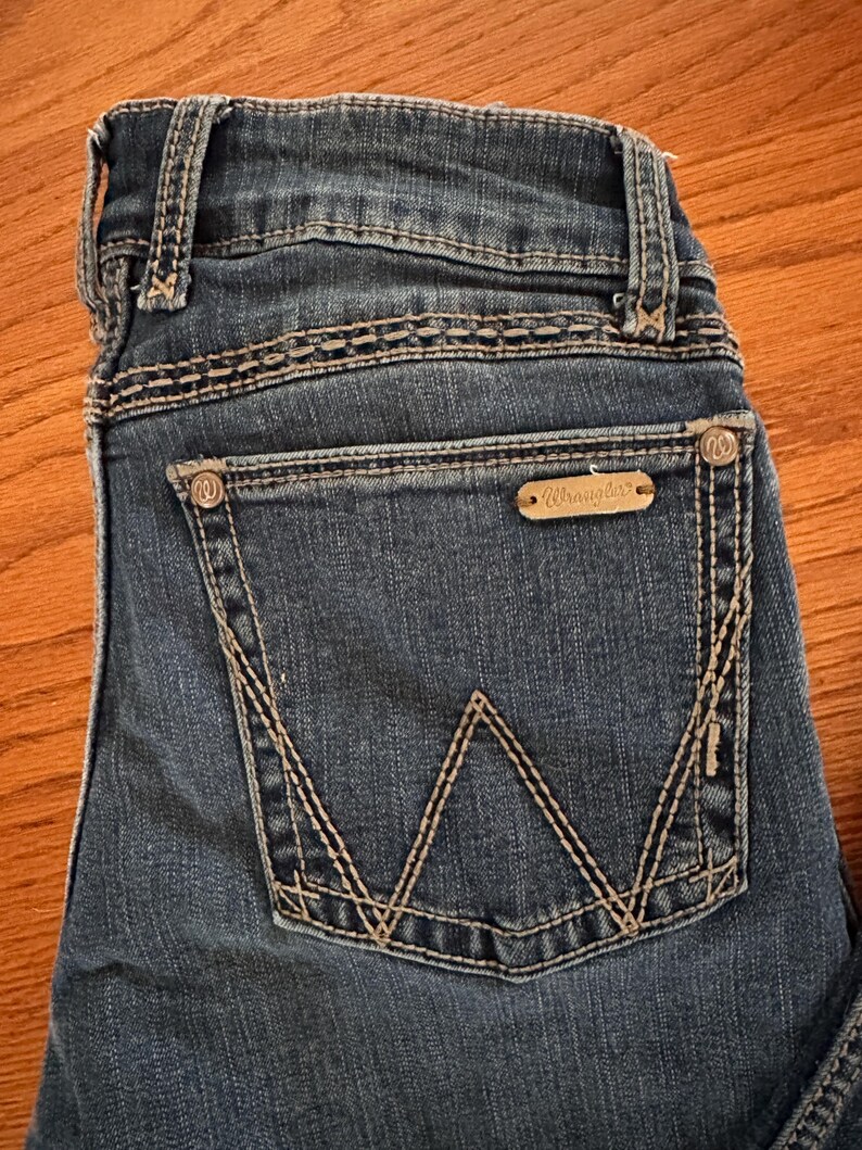 Pode incluir: Close-up do bolso traseiro de jeans azuis. O bolso apresenta um design de costura branco distinto e uma pequena etiqueta retangular com a marca "Wrangler". Os jeans t&ecirc;m um visual cl&aacute;ssico e desgastado.