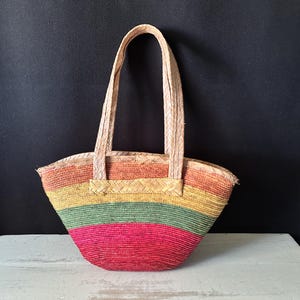 May include: A woven straw handbag featuring horizontal stripes in shades of orange, yellow, green, and pink. The bag has a curved, semi-circular base and two matching woven straw handles attached to the top edge with a braided trim detail.