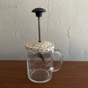 May include: A vintage glass butter churn with a white and black patterned lid and a black metal handle. The churn has markings for measuring in cups and ounces.