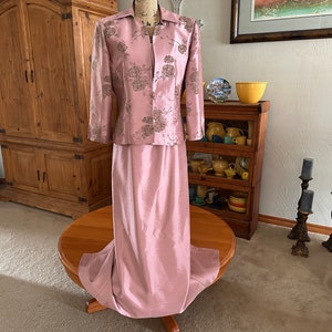 May include: A pink and silver floral patterned jacket and matching skirt. The jacket has a long sleeve and a fitted waist. The skirt is a long, flowing skirt.