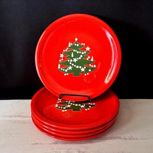 May include: A stack of red ceramic plates with a green Christmas tree design. The tree is decorated with white stars and dots. The plates are stacked on a white surface against a black background.