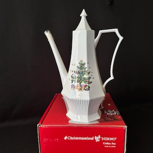 Pode incluir: Cafeteira de cerâmica branca com um desenho de árvore de Natal. A cafeteira tem um bico longo, uma alça decorativa e uma tampa pontiaguda. Está em uma caixa vermelha com as palavras "Christmastime" e "NIKKO".