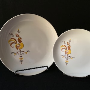 Könnte beinhalten: Zwei weiße Keramikteller mit einem Hahn-Design in Braun, Gelb und Gold. Der Hahn steht auf einem dekorativen Sockel mit einem floralen Muster.