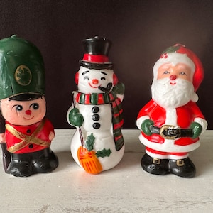 Puede incluir: Tres candelabros de Navidad vintage. Un soldado de juguete verde y rojo, un muñeco de nieve blanco con un sombrero y una bufanda verdes, y un Papá Noel rojo y blanco.