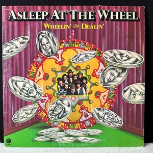 Puede incluir: Una portada de álbum de vinilo vintage con la banda "Asleep At The Wheel" con el título del álbum "Wheelin' and Dealin'". La portada presenta una rueda colorida con una foto de la banda en el centro y monedas de plata con caras que la rodean.