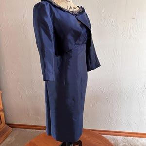 May include: A navy blue dress and jacket set displayed on a mannequin. The dress is a midi length with a fitted silhouette. The jacket has three-quarter sleeves and a fringed collar. The fabric has a subtle sheen.