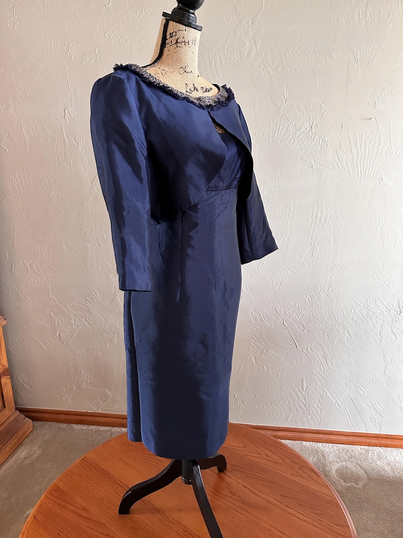 May include: A navy blue dress and jacket set displayed on a mannequin. The dress is a midi length with a fitted silhouette. The jacket has three-quarter sleeves and a fringed collar. The fabric has a subtle sheen.