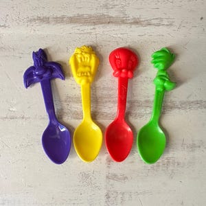 May include: Four colorful plastic spoons featuring cartoon character designs. The spoons are purple, yellow, red, and green. Each spoon has a different character design on the handle, making them a fun and playful addition to any kitchen.