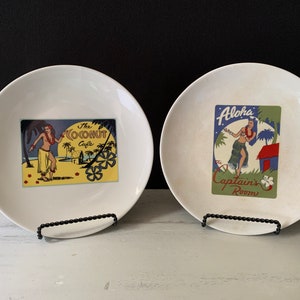 Puede incluir: Dos platos de cerámica blanca con un tema hawaiano vintage. Un plato presenta a una bailarina de hula y el texto "The Coconut Cafe". El otro plato presenta a una bailarina de hula y el texto "Aloha Captain's Room".
