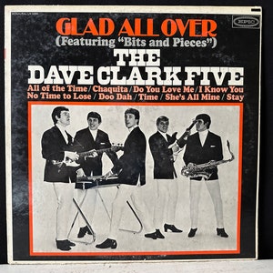 May include: A black and white vinyl record cover for the album "Glad All Over" by The Dave Clark Five. The cover features a photo of the band members in black suits and white pants, holding musical instruments. The album title is printed in red and white letters, and the band's name is printed in white letters.