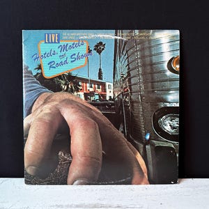 May include: A vintage vinyl record album cover featuring a hand reaching out from the bottom of the image. The album cover is titled "Hotels, Motels and Road Shows" with the band name "The Allman Brothers Band" listed below. The background features a vintage bus and a palm tree.