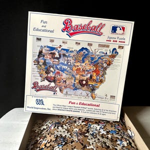 Może przedstawiać: Puzzle o tematyce baseballowej z ponad 550 elementami. Pudełko przedstawia mapę Stanów Zjednoczonych z ilustracjami związanymi z baseballem oraz słowem "Baseball" w kolorze czerwonym i niebieskim. Pudełko jest otwarte, odsłaniając elementy układanki.