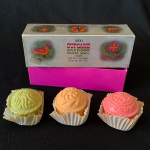 May include: A set of three Avon Cupcake Soap in a pink box. The soaps are shaped like cupcakes and are decorated with floral designs. The soaps are green, orange, and pink.