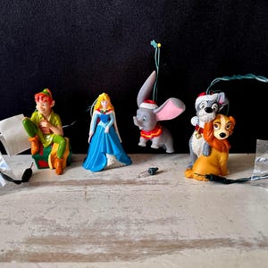 May include: A collection of four Disney character ornaments including Peter Pan sitting on a gift, Princess Aurora in a blue gown, Dumbo wearing a Santa hat, and Lady and the Tramp. Each ornament features a green light cord and small light bulbs.