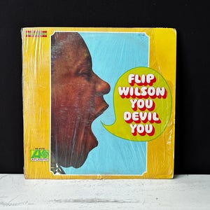 May include: A yellow vinyl record album cover with a close-up image of a person's face with their mouth open. The album title is "Flip Wilson You Devil You" in red and yellow letters.