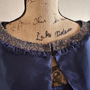 May include: A close-up of a navy blue garment with a decorative collar. The collar is embellished with sequins and a fringed edge. The garment has a simple, open front design.