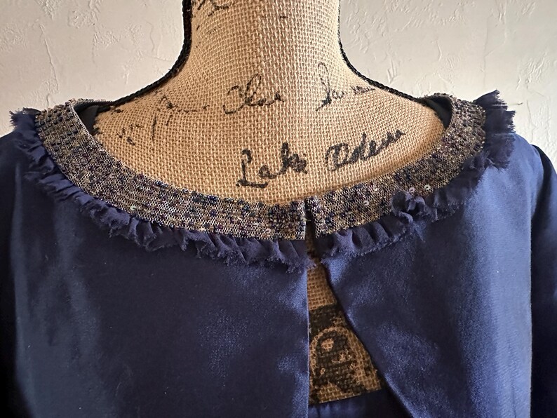 May include: A close-up of a navy blue garment with a decorative collar. The collar is embellished with sequins and a fringed edge. The garment has a simple, open front design.