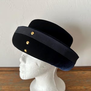Puede incluir: Un sombrero de terciopelo negro con una cinta azul oscuro y tachuelas doradas. El sombrero se exhibe en una cabeza de maniquí blanca. Tiene una copa redondeada y un ala plana, de estilo vintage o retro.