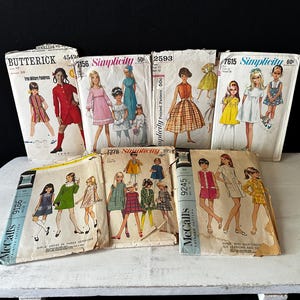 May include: A collection of vintage sewing patterns for girls' dresses. The patterns are from the 1960s and feature a variety of styles, including A-line dresses, shift dresses, and dresses with Peter Pan collars. The patterns are in various colours, including red, blue, green, and yellow.