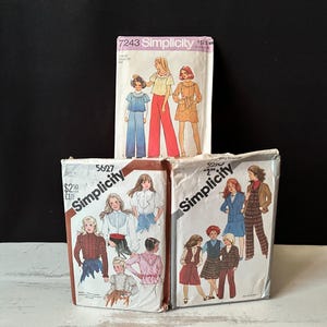 May include: Three vintage Simplicity sewing pattern packets. The packets feature illustrations of women's and children's clothing designs. The packets are stacked on a white surface against a black background.