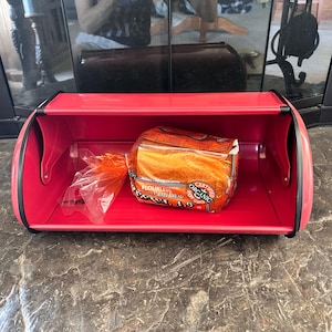 Op de afbeelding: Een rode metalen broodtrommel met een brood van biologisch brood erin. Het brood zit in een doorzichtige plastic zak. De broodtrommel heeft een gebogen bovenkant en zijkanten en een zwarte rand.