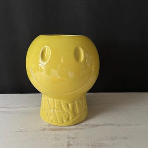 Pode incluir: Um vaso amarelo de cerâmica com um rosto sorridente, com uma cabeça redonda e uma base que diz "HAVE A HAPPY DAY". O vaso tem um acabamento brilhante e está sobre uma superfície branca.