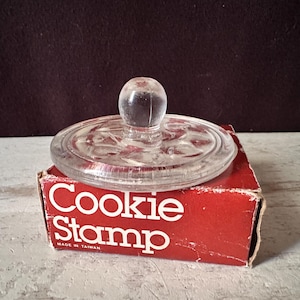 May include: A clear glass cookie stamp with a round handle sits atop a red cardboard box. The box features white text that reads Cookie Stamp and Made in Taiwan. The stamp has a circular base with a molded pattern on the underside surface.