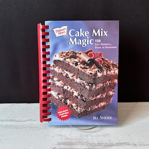 May include: A cookbook titled "Cake Mix Magic" by Jill Snider, featuring a layered chocolate cake with frosting and toppings. The book has a red spiral binding and a blue cover with the Duncan Hines logo.